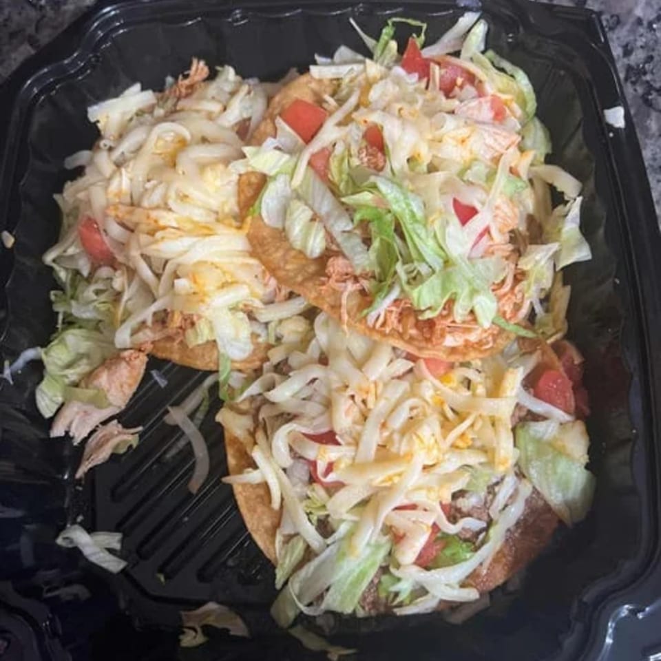 Three Dos Pesos Mexican Restaurant tostadas with shredded chicken, lettuce, tomatoes, and cheese in a black container.