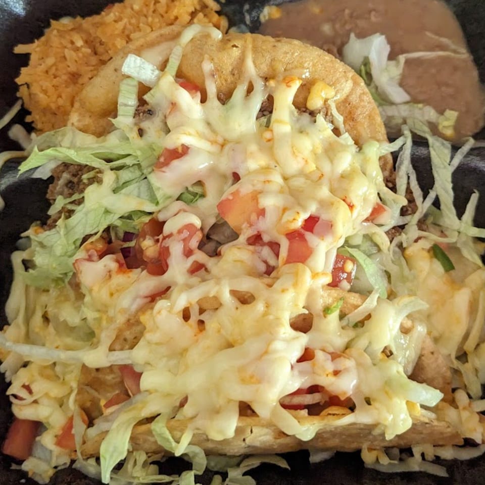Two Dos Pesos Mexican Restaurant tacos topped with melted cheese, lettuce, and diced tomatoes, with rice and beans.