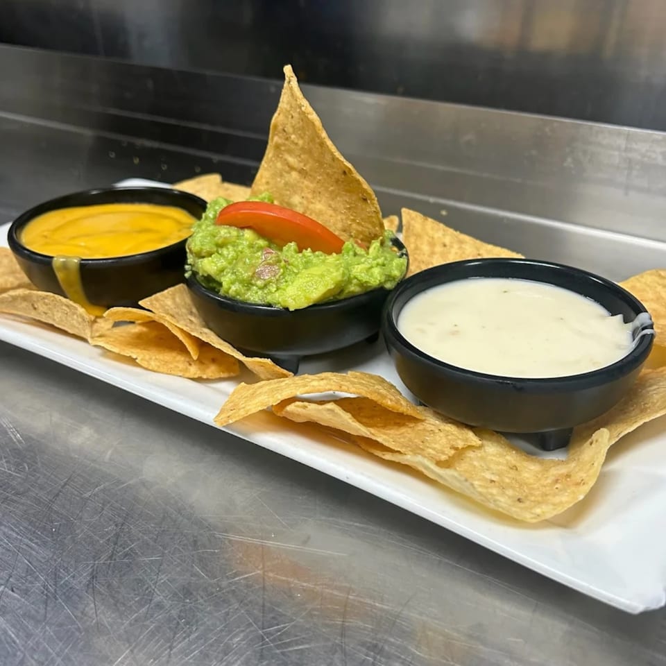 A white platter of tortilla chips with three bowls of dips: queso, guacamole, and white cheese dip.